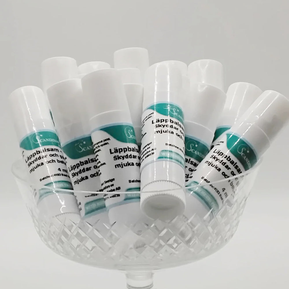 A glass dish holds several upright tubes of Läppbalsam - för alla årstider by Scandinavian Herbs. Each tube has a white cap and a teal and white Swedish label listing ingredients like sheasmör and vegetabiliska oljor, with some tubes artistically tilted.