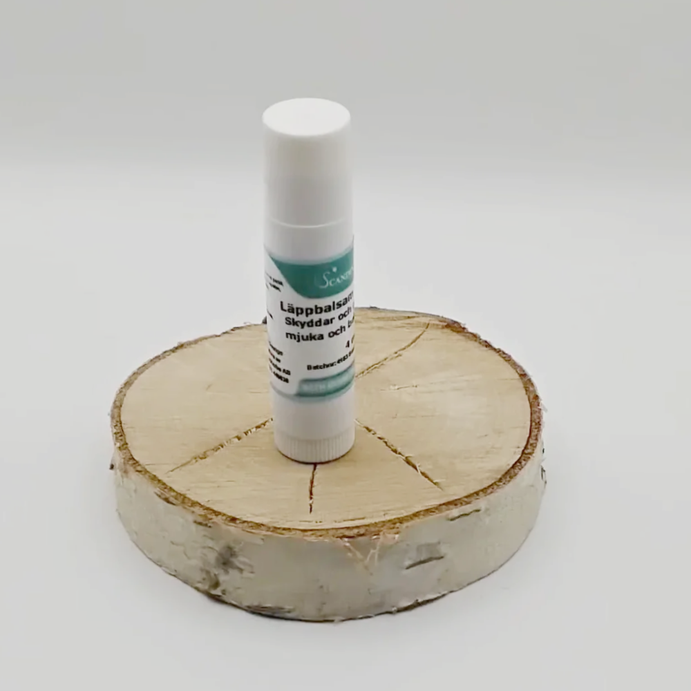 A tube of Scandinavian Herbs Läppbalsam - för alla årstider sits gracefully on a circular wooden slice, its blue label striking against the plain white background. Enriched with sheasmör and vegetabiliska oljor, it provides ultimate nourishment.