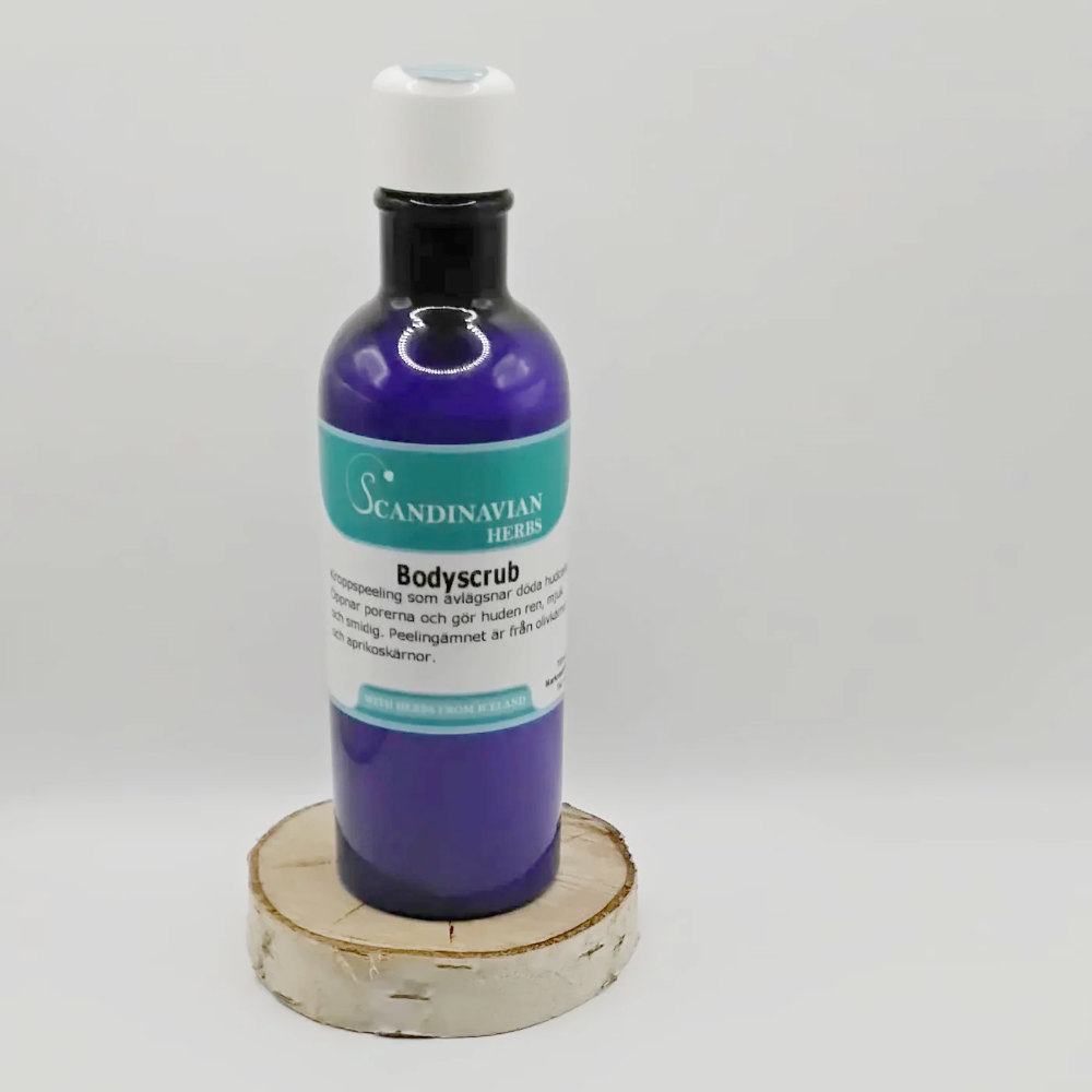 A purple bottle of Scandinavian Herbs Bodyscrub - härlig scrub för hela kroppen with olivkärnor rests on a wooden round base. It has a pump dispenser, plus a green and white label displaying product details against a simple white backdrop.