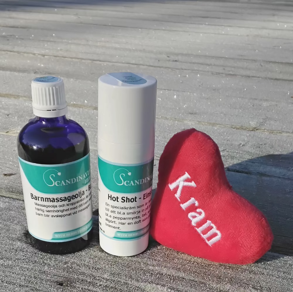 Two bottles labeled Scandinav sit on a wooden surface beside a small red fabric heart with Kram. A bottle of barnmassageolja glistens nearby. In the background, sunlight casts shadows on the wood. This scene encapsulates the essence of Dagispaket by Örtkraft, perfect for small children and parents.