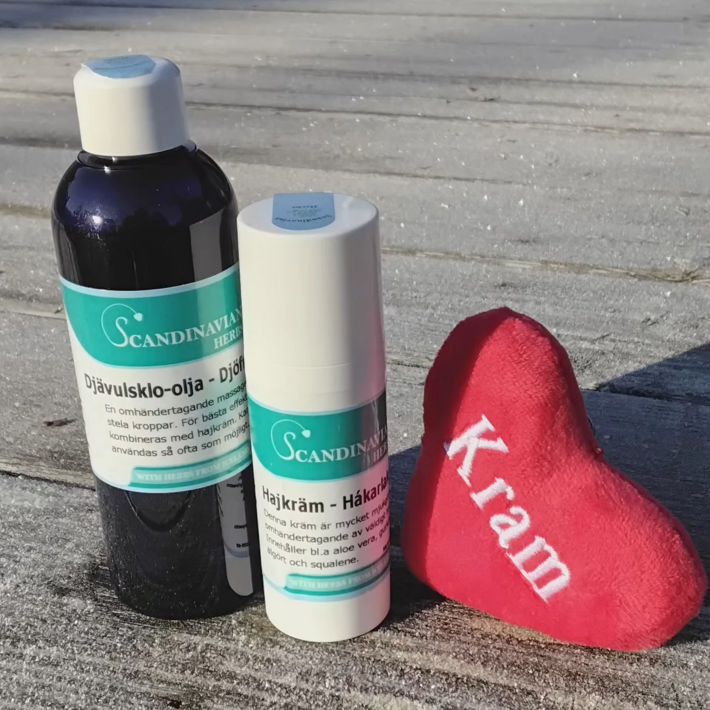 A Friskvårdspaket - för stela och ömma leder by Scandinavian Herbs rests on a frosty wooden deck, accompanied by a small red plush heart with Kram. A building looms in the background.