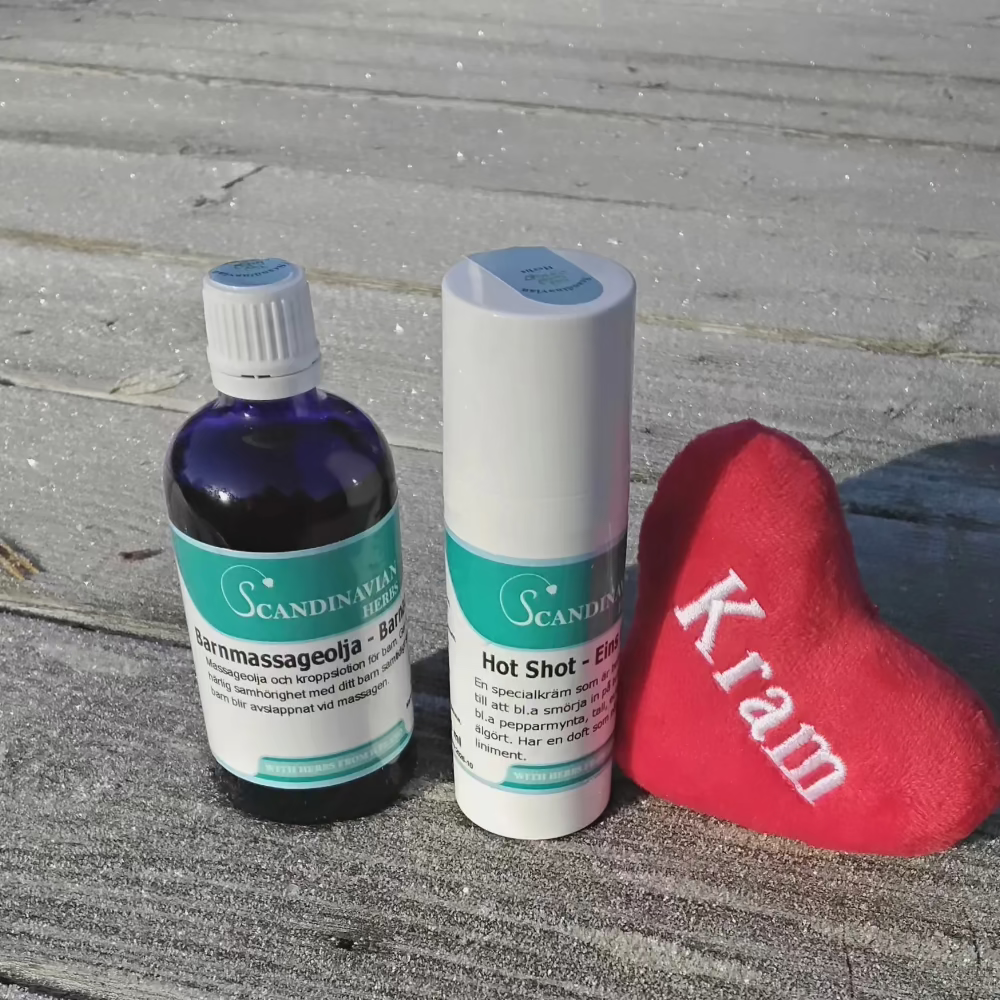 On a frosty wooden surface, a blue bottle, a white roll-on stick labeled Hot Shot Gel, and a small red heart embroidered with Kram rest. To the right, theres Barnmassageolja for soothing comfort. All items are part of the Dagispaket - för alla småbarnsföräldrar by Örtkraft.