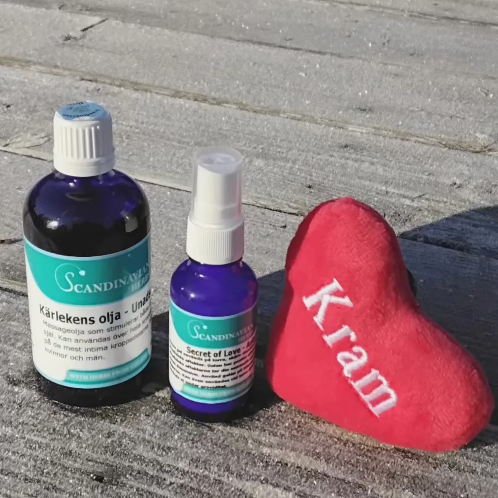 Two blue bottles labeled in Scandinavian text, hinting at the Secret of Love, and a red heart-shaped cushion with Kram embroidered, are set on a wooden surface under sunlight. The Kärlekspaket- en relationshöjare by Scandinavian Herbs includes bottles of different sizes.