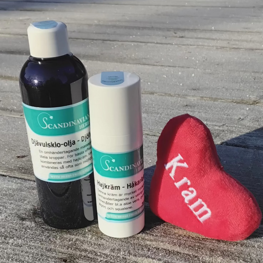A Friskvårdspaket - för stela och ömma leder by Scandinavian Herbs rests on a frosty wooden deck, accompanied by a small red plush heart with Kram. A building looms in the background.
